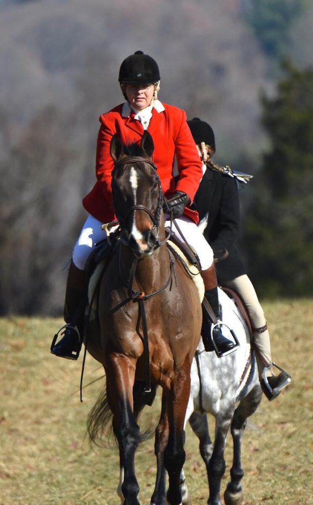 Woman in red coat hunting on a dark horse, followed by a girl on a gray pony